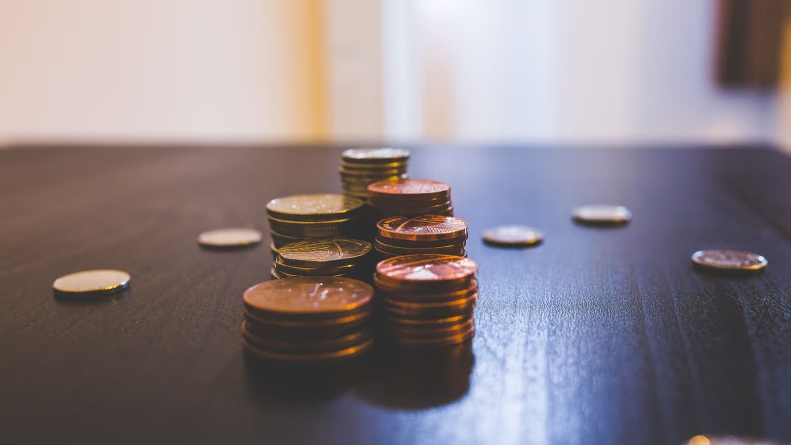 Petites piles de pièces de centimes posées sur une table en bois sombre, quelques pièces éparses tout autour, dans la lumière chaude d'un intérieur, image de la monnaie du quotidien mise de côté