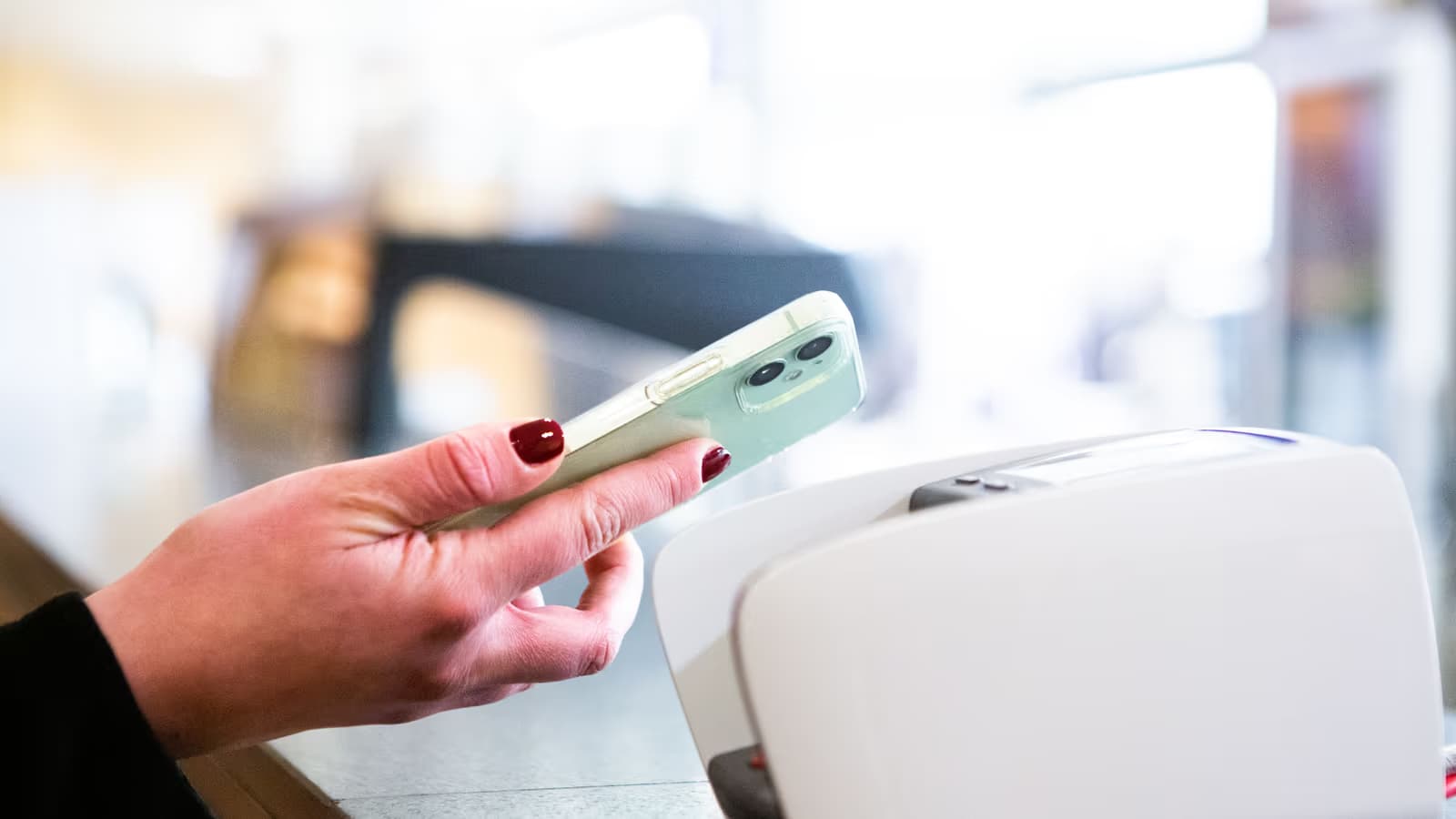 Main tenant un téléphone au-dessus d'un terminal de paiement sans contact blanc, sur le comptoir clair d'un commerce, geste quotidien du paiement mobile