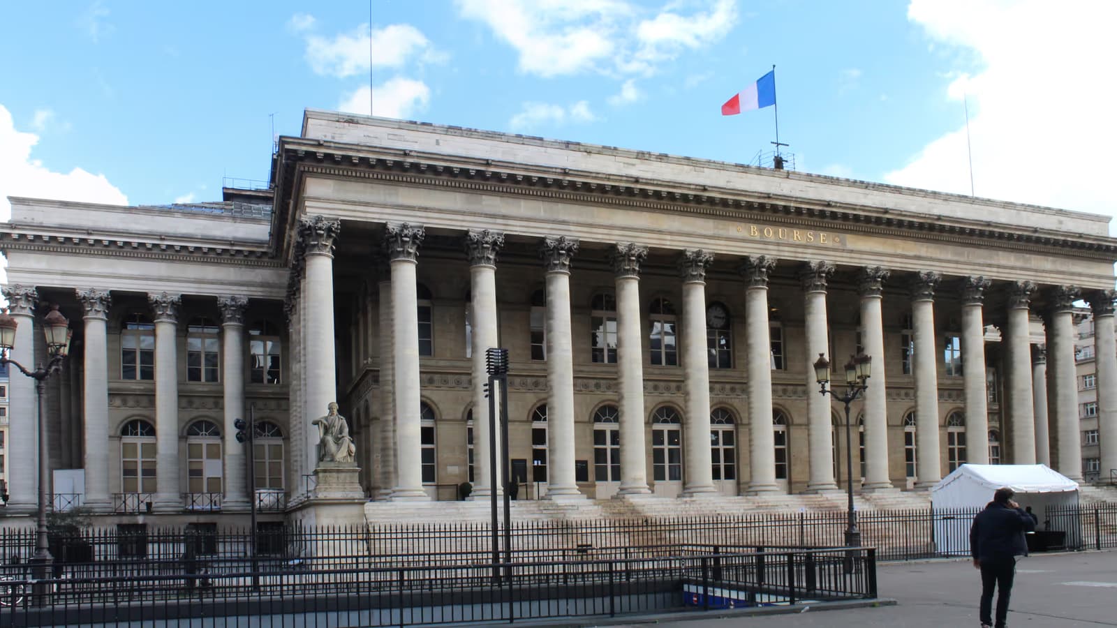 Façade à colonnades du Palais Brongniart à Paris, ancien palais de la Bourse, surmontée du drapeau français sous un ciel bleu nuageux