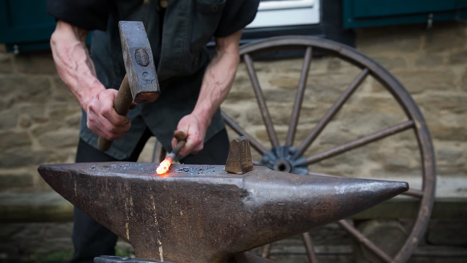 Forgeron frappant au marteau une barre de métal incandescente posée sur une enclume, devant un mur de pierre et une roue de charrette, image du travail quotidien d'un petit atelier artisanal