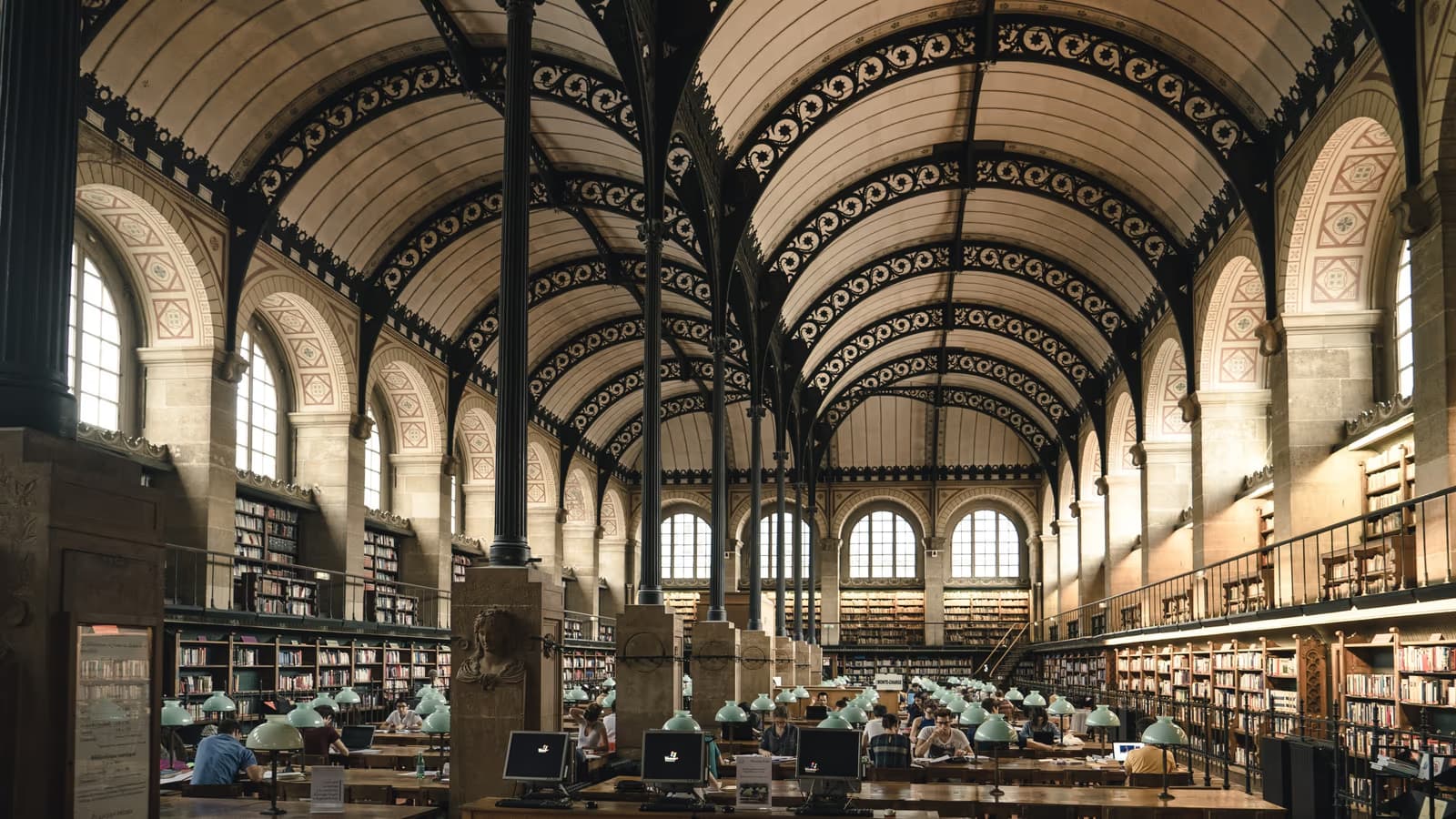 Grande salle de lecture voûtée de la bibliothèque Sainte-Geneviève à Paris, lecteurs installés aux longues tables sous les lampes vertes, entourés de rayonnages de livres jusqu'au plafond
