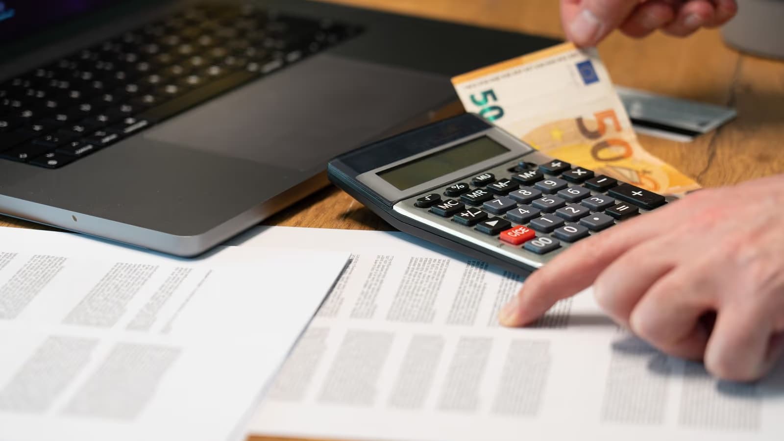 Personne pointant du doigt un document financier près d'une calculatrice, d'un ordinateur portable et d'un billet de 50 euros posés sur un bureau en bois, scène de calcul pour renégocier et optimiser le coût de son crédit