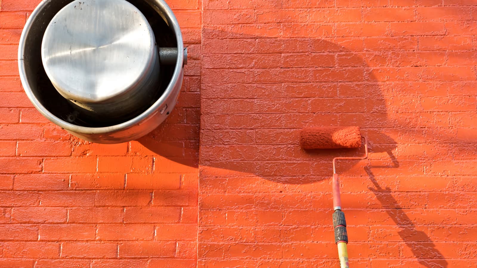 Rouleau appliquant une peinture terracotta sur un mur de briques en cours de rénovation, à côté d'un pot de peinture métallique