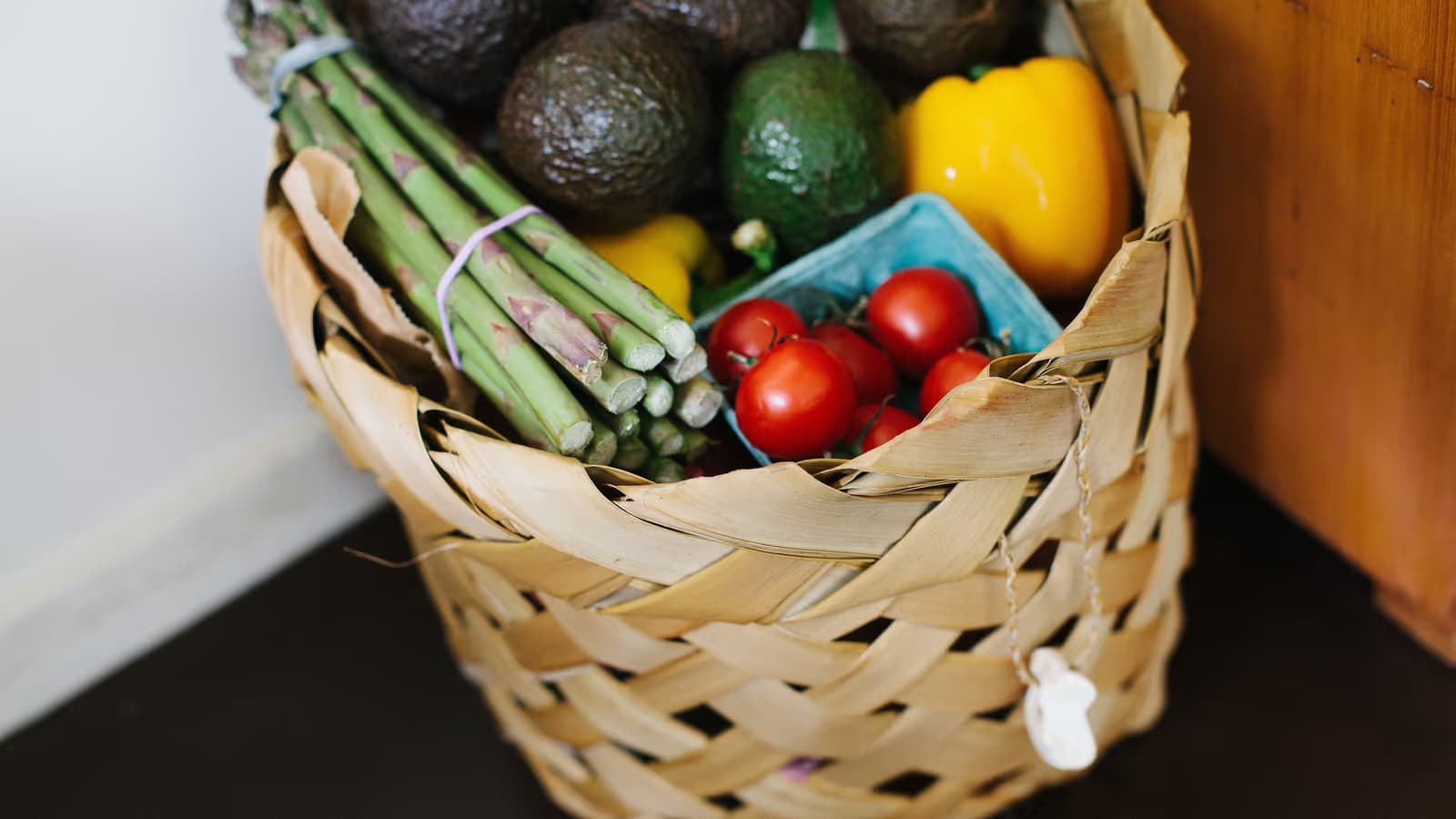 Panier en osier rempli de légumes frais du marché — asperges, avocats, poivron jaune et tomates cerises — posé au pied d'un meuble en bois, scène de courses quotidiennes faites avec soin